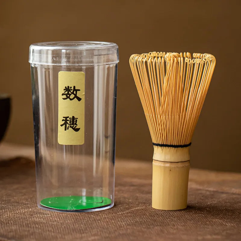 Bamboo chasen (matcha whisk) with handwoven bristles and a black twine-wrapped handle, paired with a clear plastic storage case featuring a gold label inscribed with Japanese characters ' 数穂 ', presented on a brown textured surface for traditional Japanese tea ceremonies.