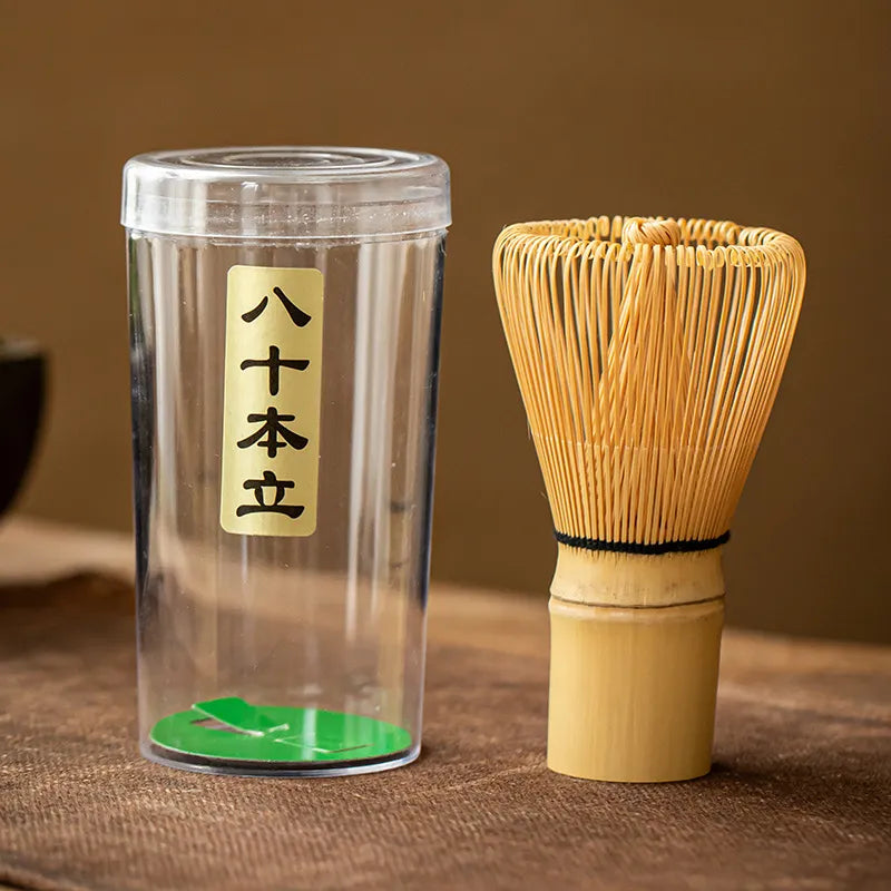 Bamboo chasen (matcha whisk) with an 80 - prong design, paired with a clear plastic storage case featuring a gold label inscribed with Japanese characters ' 八十本立 ' (Hajihon Dachi), displayed on a brown textured surface for traditional Japanese tea ceremonies.