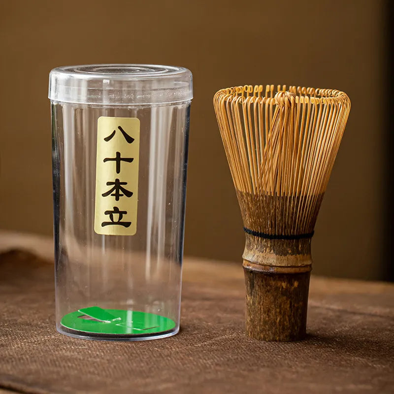 Bamboo chasen (matcha whisk) crafted from dark Zizhu bamboo, with handwoven bristles and a black twine-wrapped handle, paired with a clear plastic storage case featuring a gold label inscribed ' 八十本立 ' (Hajihon Dachi, indicating 80-prong design), displayed on a brown textured surface for traditional Japanese tea ceremonies.