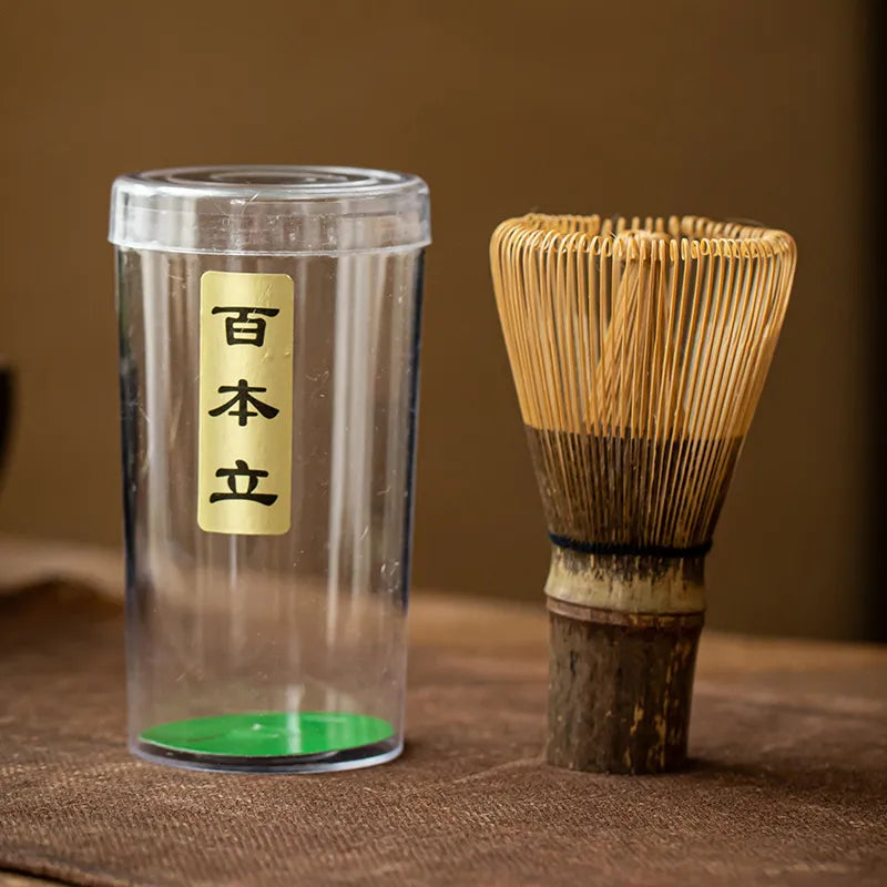 Bamboo chasen (matcha whisk) crafted from dark Zizhu bamboo, featuring handwoven bristles, a black twine-wrapped handle, and paired with a clear plastic storage case with a gold label inscribed ' 百本立 ' (Hyakubon Dachi, indicating 100-prong design), displayed on a brown textured surface for traditional Japanese tea ceremonies.