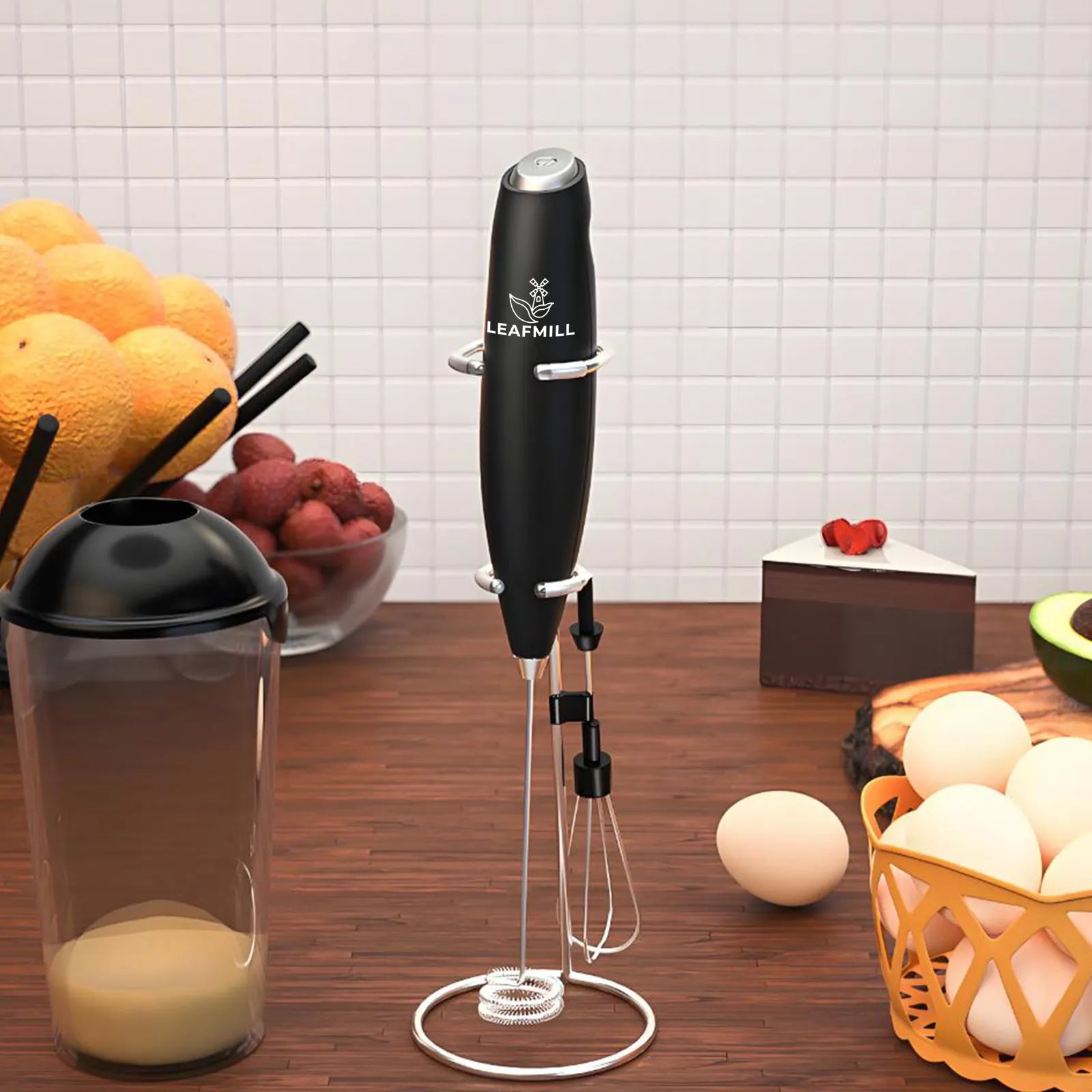 Leafmill electric matcha frother in black with stand and dual whisk attachments, placed on a kitchen counter with eggs, fruit, a dessert slice, and a mixing cup nearby