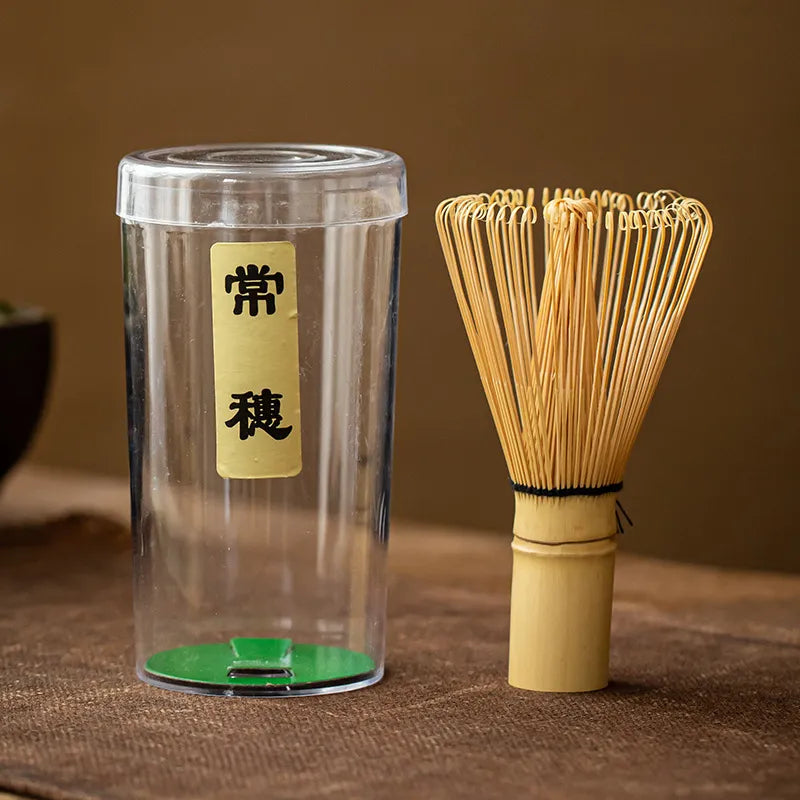 Bamboo chasen (matcha whisk) with handwoven bristles and a black twine-wrapped handle, paired with a clear plastic storage case featuring a gold label inscribed with Japanese characters ' 常穗 ', displayed on a brown textured surface for traditional Japanese tea ceremonies.