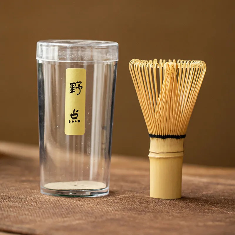 Bamboo chasen (matcha whisk) with a black twine-wrapped handle, paired with a clear plastic storage case featuring a gold label inscribed with Japanese characters ' 野点 ', displayed on a brown textured surface for traditional Japanese tea ceremonies.